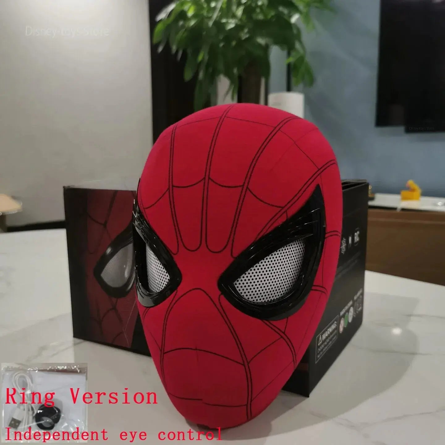 Spider mask with black mesh eye covers, red fabric with black web pattern, displayed on a table with a plant and box in the background.